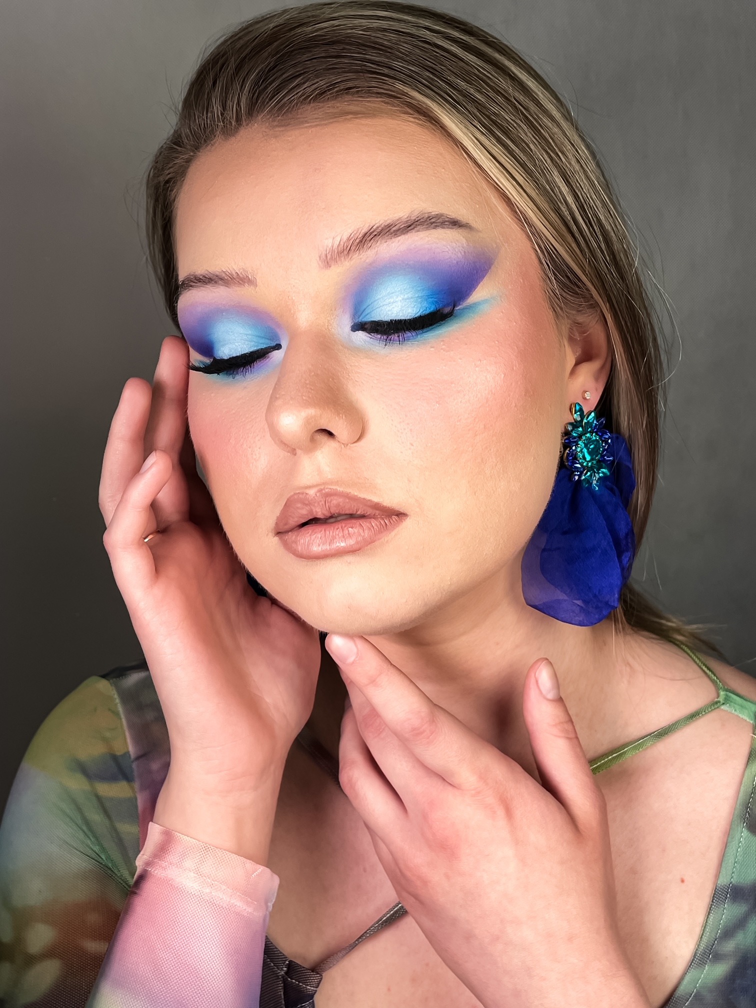 Portret kobiety z zamkniętymi oczami, prezentującej fantazyjny, niebiesko-fioletowy makijaż oczu, duże, niebieskie kolczyki z materiału i delikatny makijaż ust.