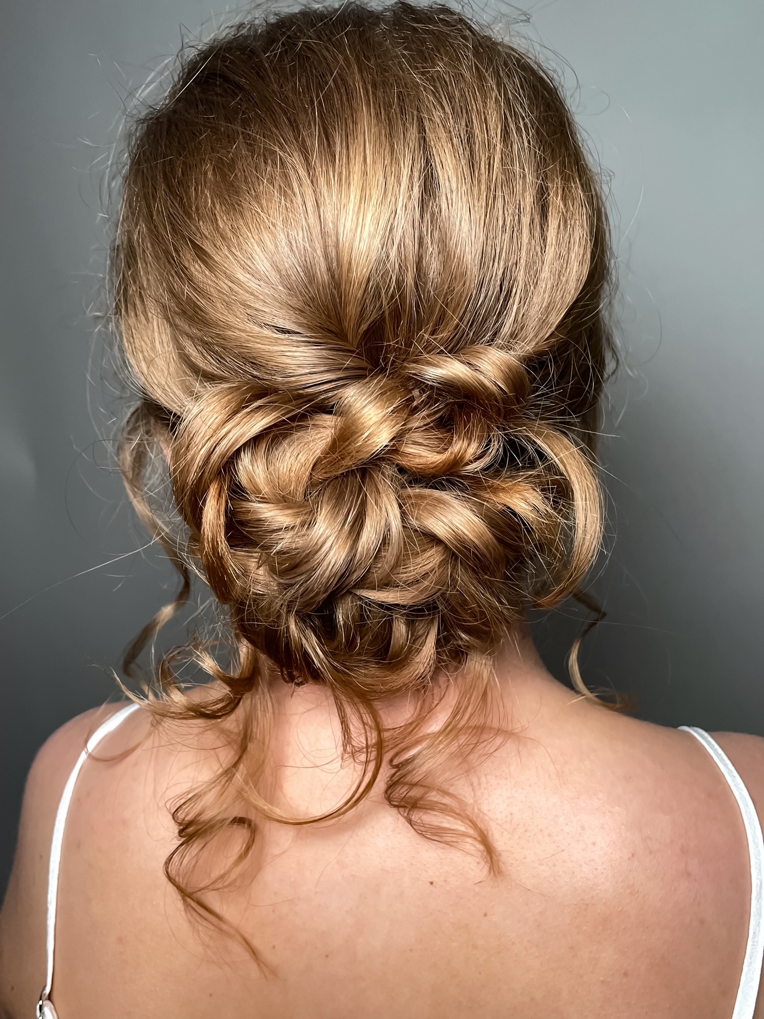 Upięcie ślubne z blond włosów, kok z loków z tyłu głowy, widoczne pojedyncze kosmyki opadające na kark.