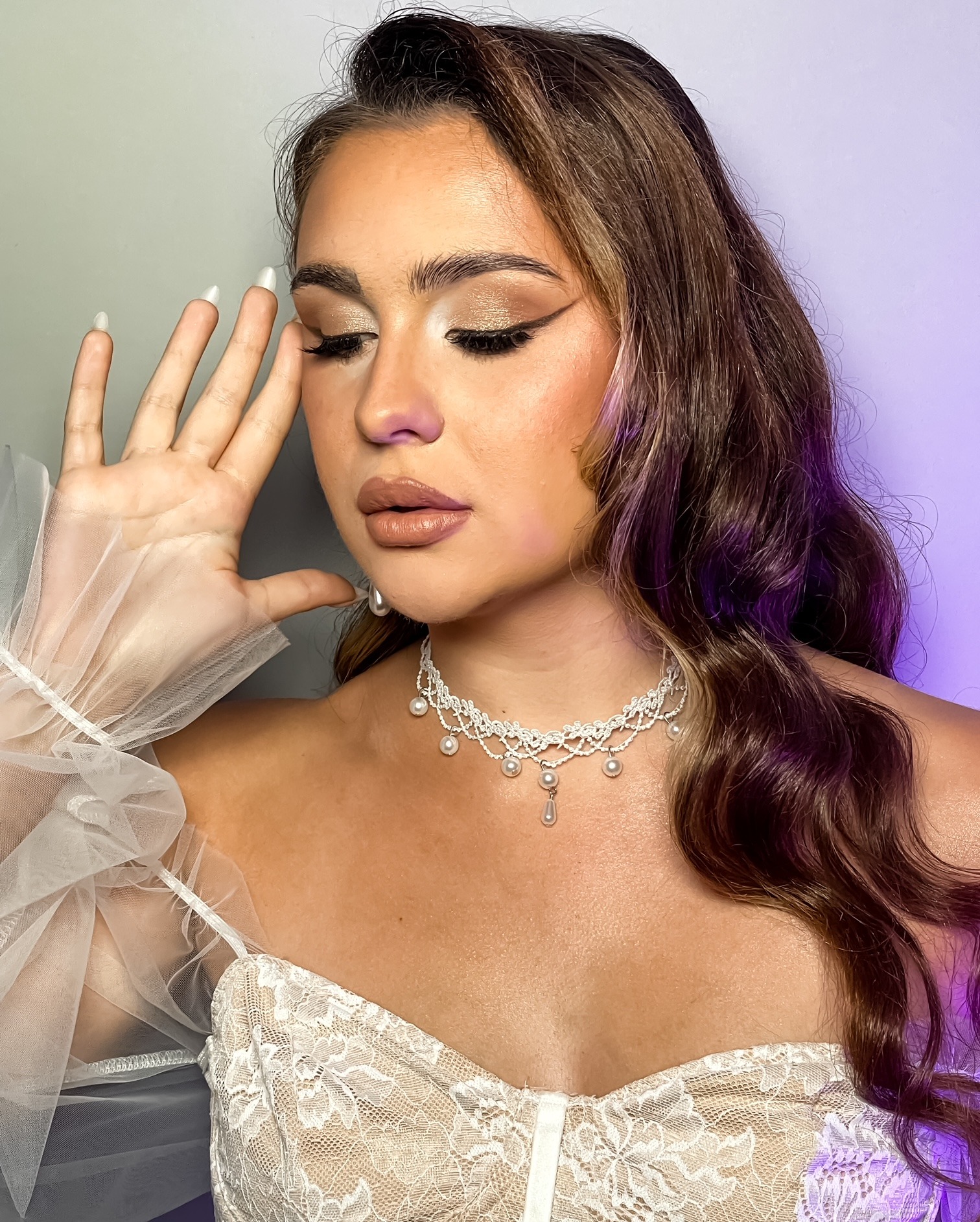 Portret kobiety z profesjonalnym makijażem ślubnym w odcieniach złota, z delikatną biżuterią z pereł i koronkową sukienką, pozującej z zamkniętymi oczami i dłonią przy twarzy na tle z fioletowym...