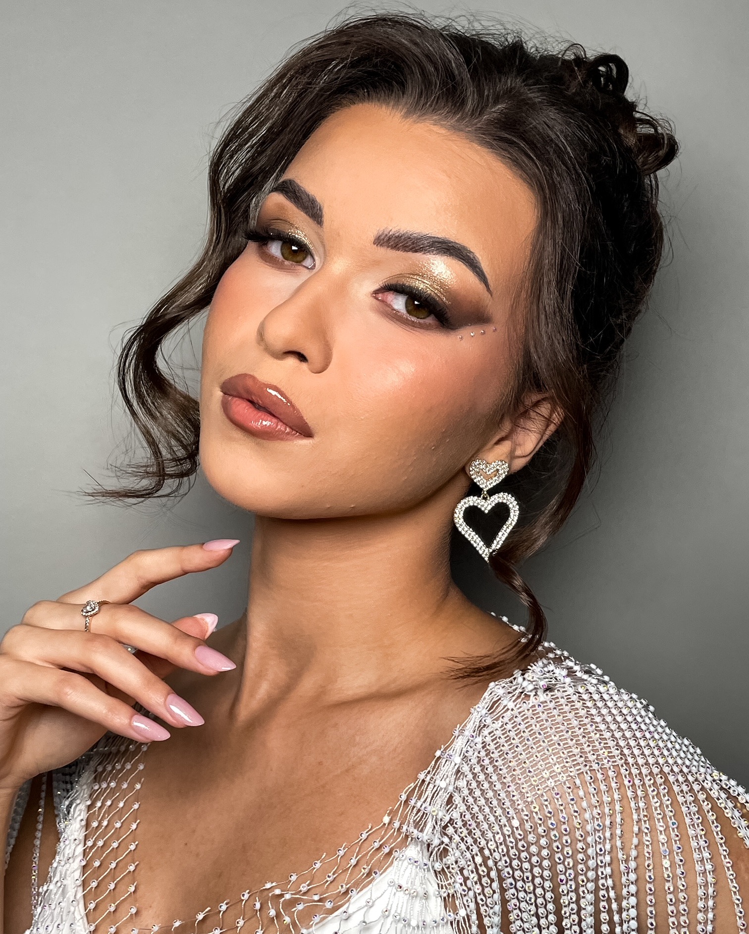 Portret kobiety z błyszczącym, złotym cieniem do powiek, drobnymi kryształkami pod okiem, błyszczącymi ustami, upiętymi ciemnymi włosami z wypuszczonymi lokami, w ozdobnej kreacji z koralików i...