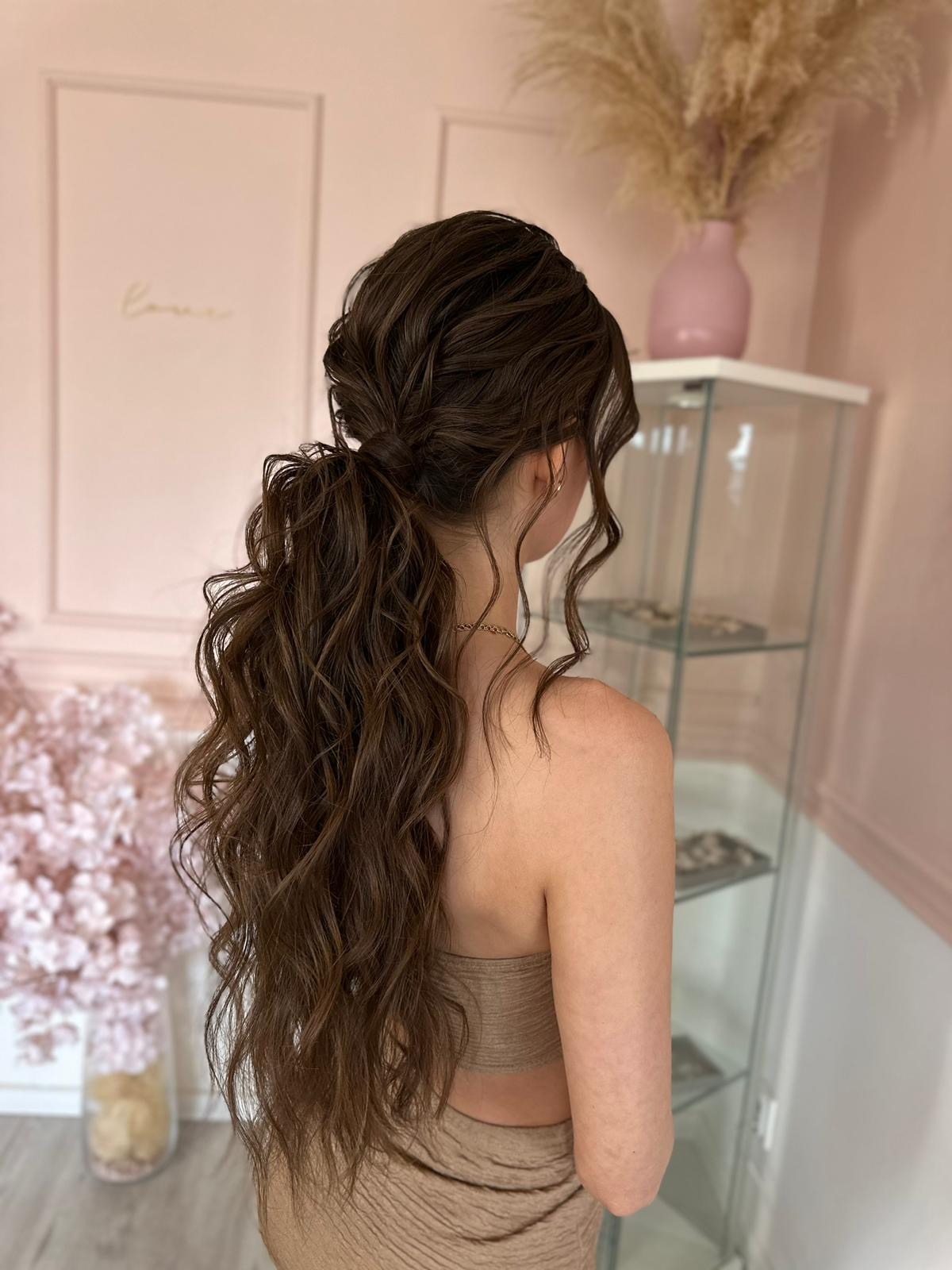 Elegancka fryzura: upięcie z brązowych, falowanych włosów, związanych w niski kucyk. Widok z tyłu, na jasnym tle z dekoracjami. Subtelna biżuteria na szyi.