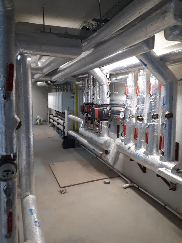 Izolowane rury instalacji grzewczej w kotłowni, widoczne zawory, manometry i centrala sterująca.
