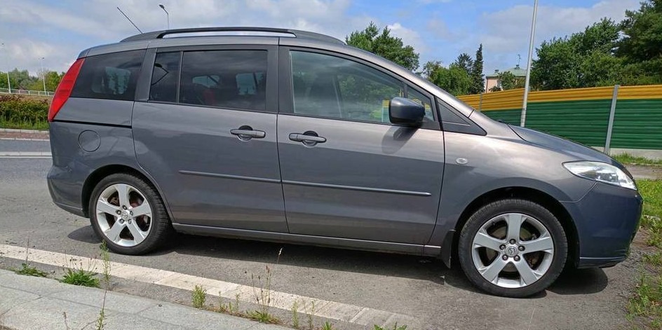 Szary minivan Mazda 5 zaparkowany na poboczu drogi, z widocznymi detalami felg i relingów dachowych.