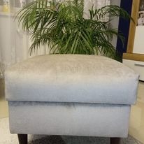 Pufa tapicerowana w odcieniach szarości, z widocznymi ciemnymi nóżkami, na tle rośliny doniczkowej i mebli w tle. Delikatne oświetlenie.