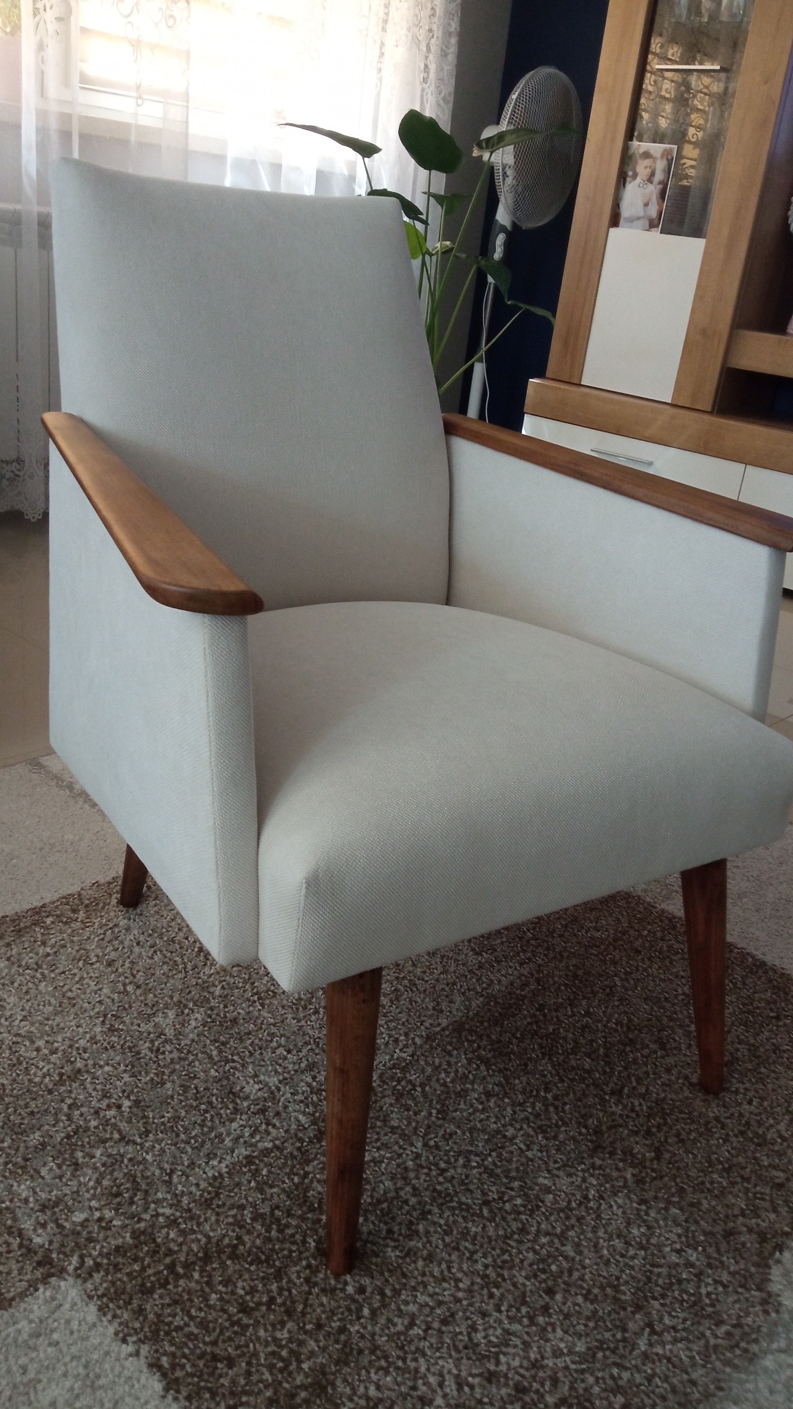 Biały fotel z drewnianymi podłokietnikami i nogami, stojący na brązowym dywanie. W tle okno z firanką, roślina i meble.