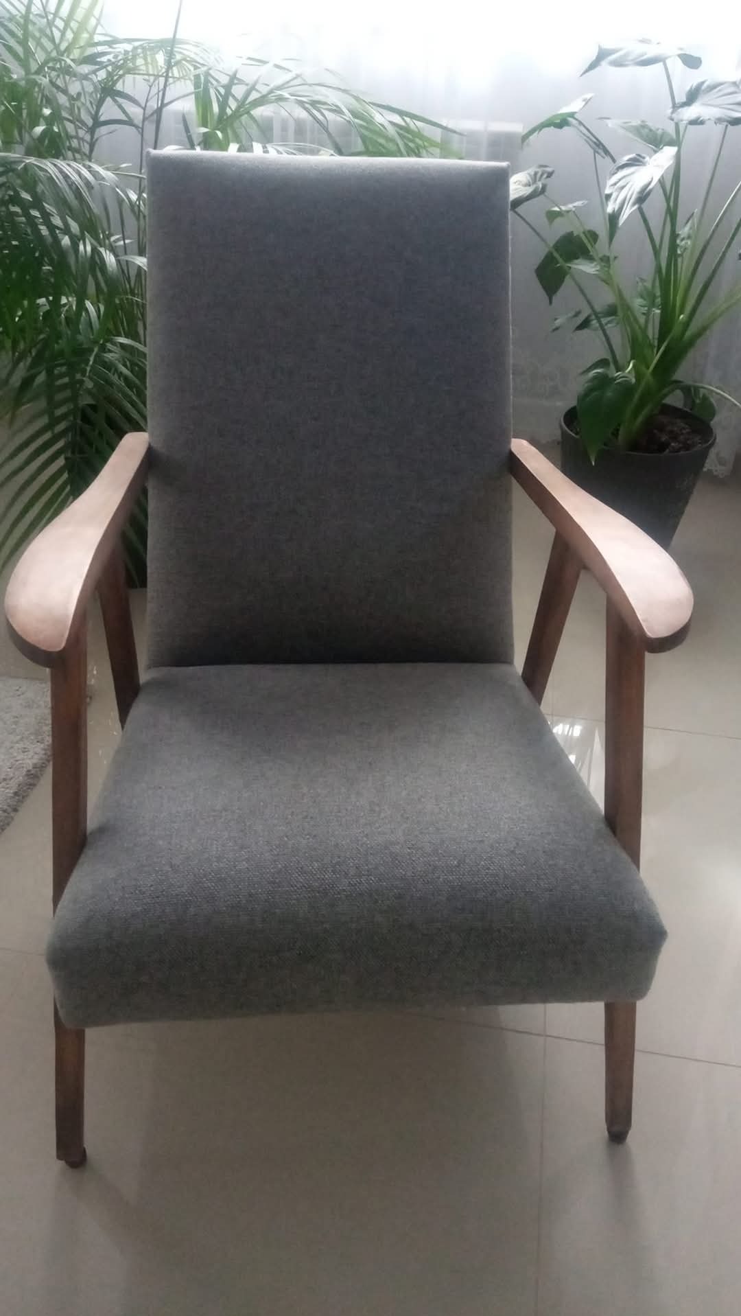 Odnowiony fotel w szarej tapicerce z drewnianymi podłokietnikami, na jasnej podłodze, z roślinami w tle. Usługa tapicerska.