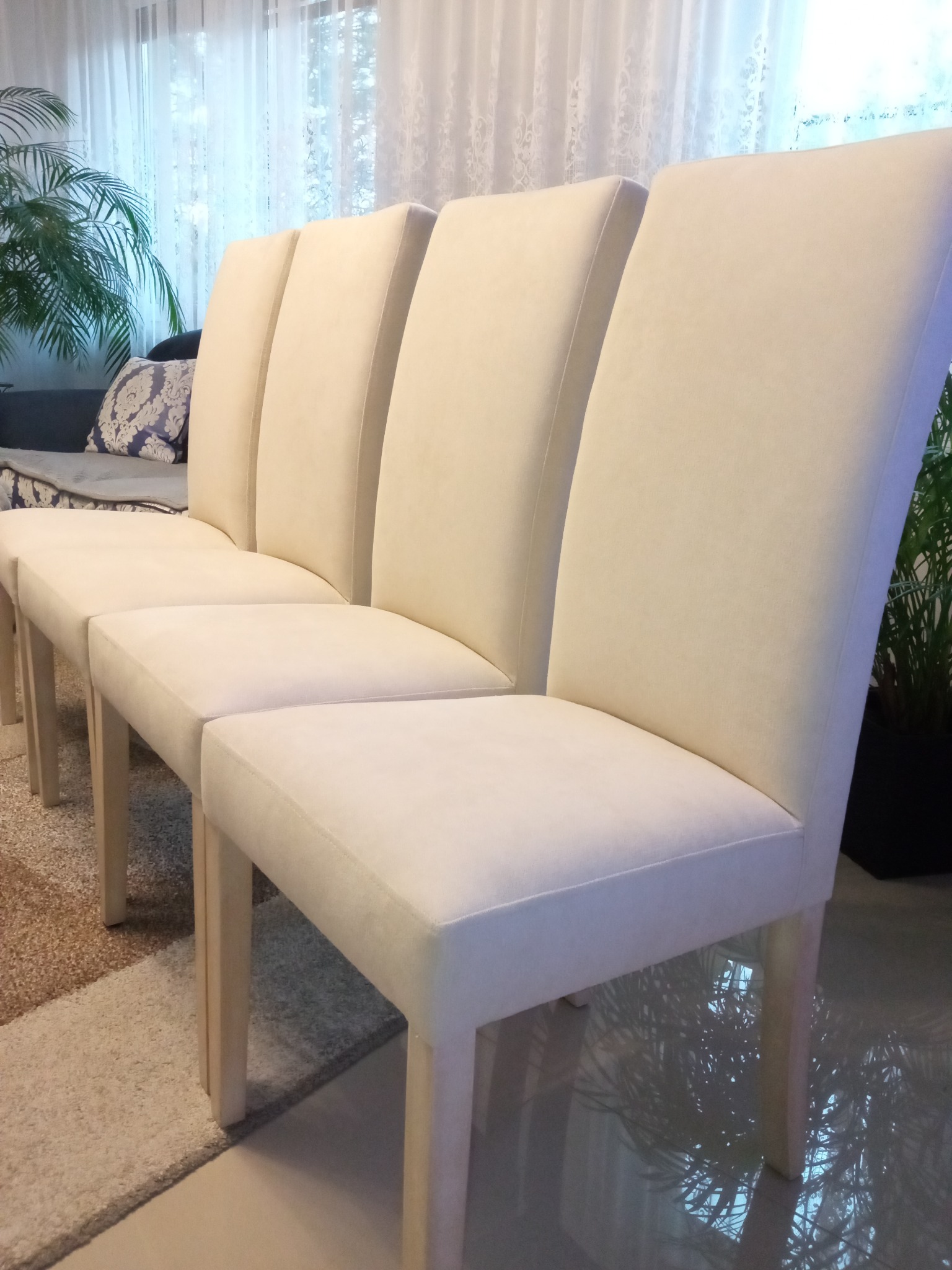 Rząd czterech krzeseł z wysokim oparciem, obitych kremową tkaniną, ustawionych na tle salonu z roślinami doniczkowymi i wzorzystym dywanem.