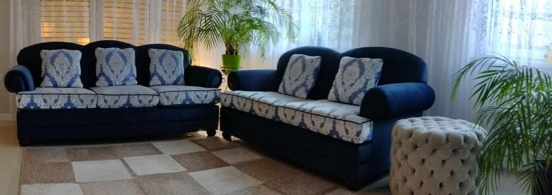 Komplet wypoczynkowy w odcieniach granatu i beżu z wzorzystymi poduszkami, uzupełniony pufem i rośliną doniczkową na tle okna z firankami.