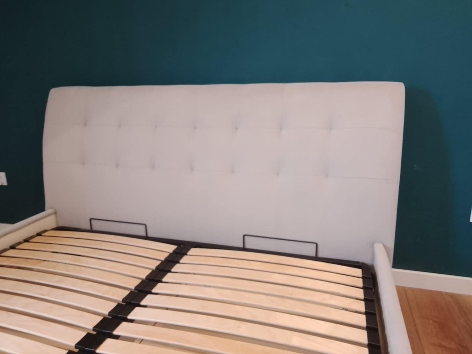 Tapicerowane wezgłowie łóżka w jasnym odcieniu z subtelnymi przeszyciami, widoczny stelaż z drewnianych listew na tle ciemnozielonej ściany.