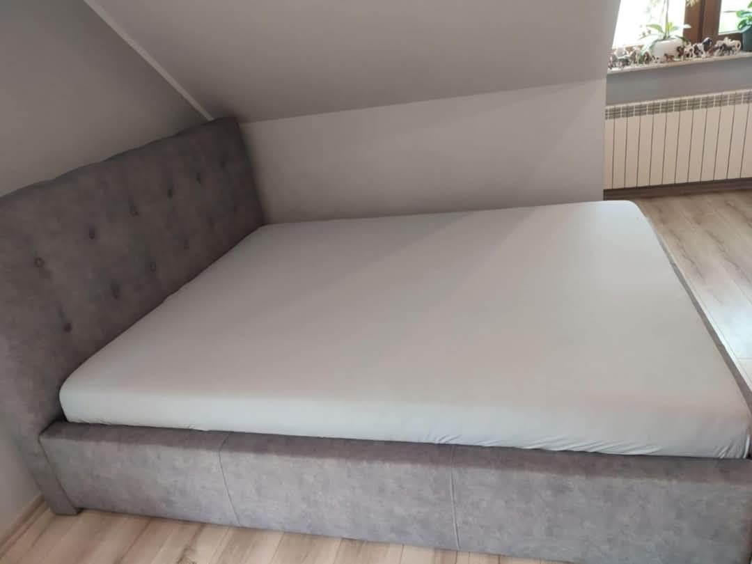 Szare, tapicerowane łóżko małżeńskie z zagłówkiem w pokoju na poddaszu, nakryte białym prześcieradłem. Widoczny fragment okna i grzejnika.