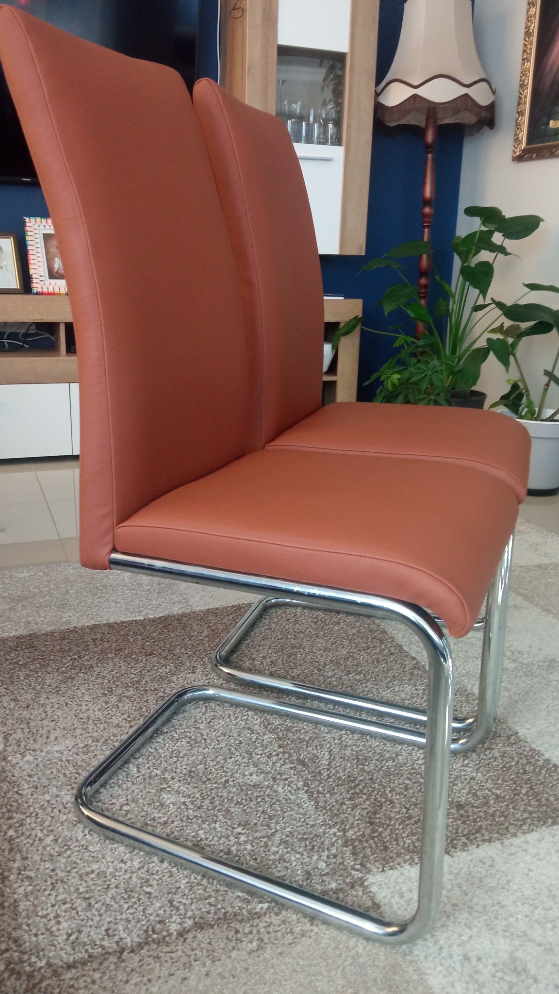 Brązowe krzesło z chromowanymi nogami w kształcie sanek, na tle dywanu i mebli. Widok z boku, oświetlenie dzienne. Krzesło po renowacji.