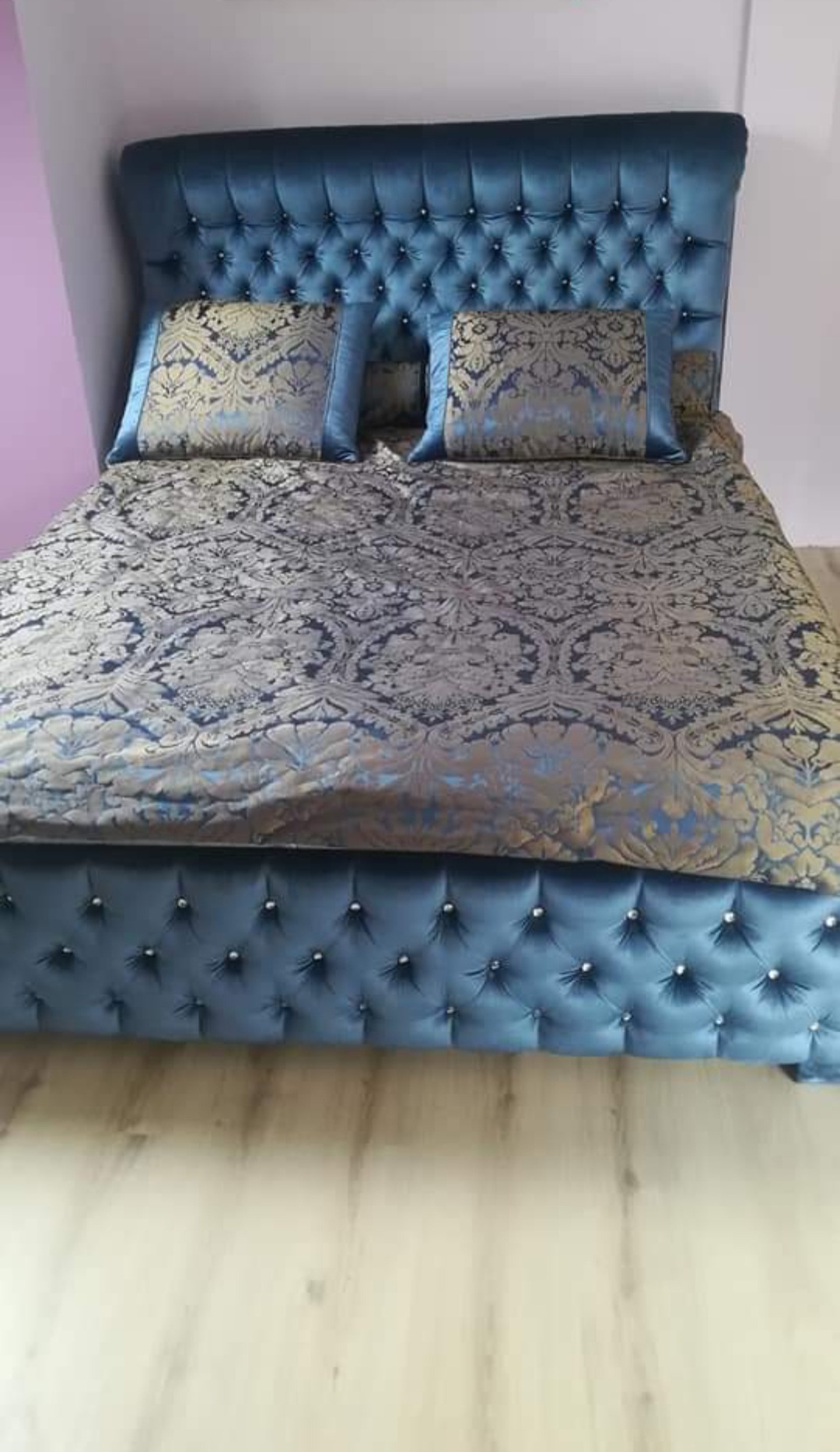 Niebieskie, tapicerowane łóżko z pikowanym zagłówkiem i wzorzystą pościelą, widoczne na jasnej podłodze. Dekoracyjne guziki na ramie i zagłówku.