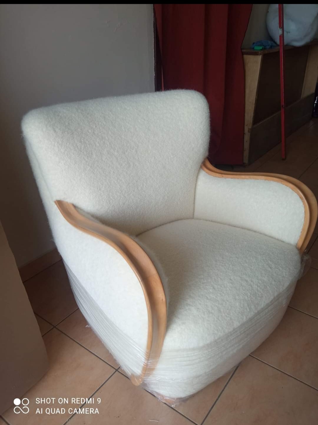 Kremowy fotel z drewnianymi podłokietnikami, zabezpieczony folią stretch, stoi na brązowej podłodze. Widoczne tło z zasłoną i meblami.