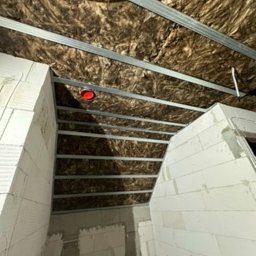 Mateusz brzeń - Sufit poddasza z widoczną wełną mineralną między metalowymi profilami stelaża, przygotowany pod zabudowę płytami, z fragmentami ścian z białych bloczków.