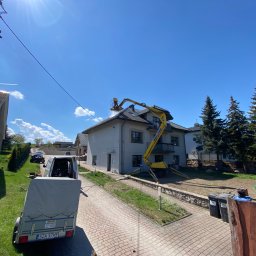 Czysta Strefa Mateusz Wójcik - Żółty podnośnik koszowy przy szarym domu podczas mycia dachu. Widoczny fragment kostki brukowej, trawnik, drzewa iglaste i przyczepa. Słoneczny dzień.