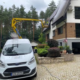 Czysta Strefa Mateusz Wójcik - Pracownik na podnośniku czyści dach domu z drewnianą elewacją. Biały van z logo firmy na pierwszym planie, otoczony zielenią i kostką brukową.