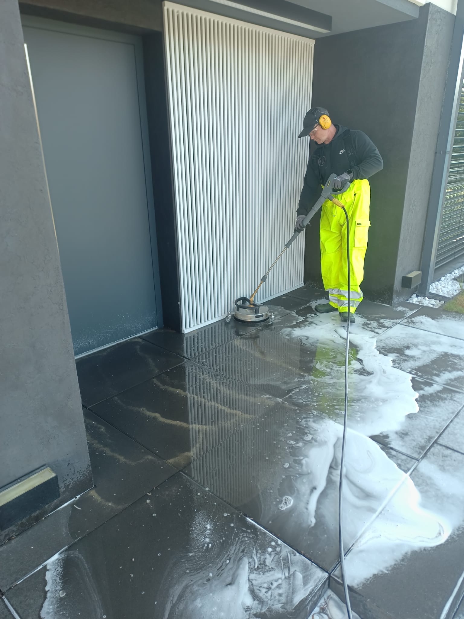 Pracownik w odzieży ochronnej czyści ciemną kostkę brukową przed budynkiem za pomocą myjki ciśnieniowej. Widoczne białe pozostałości po czyszczeniu.