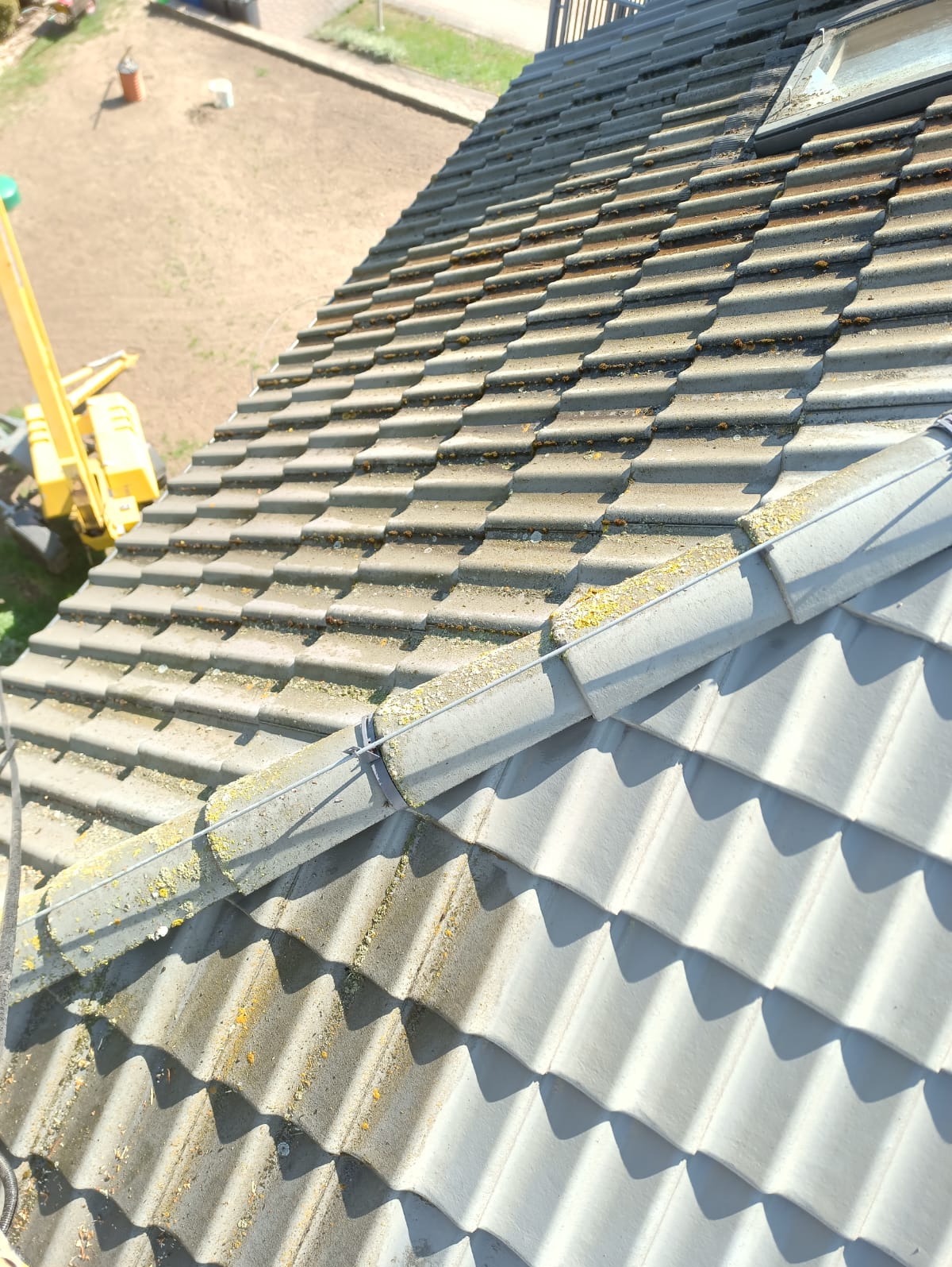 Częściowy widok na dachówkę z mchem i zabrudzeniami, z żółtym podnośnikiem w tle. Fragment dachu po czyszczeniu, ukazujący różnicę w kolorze.