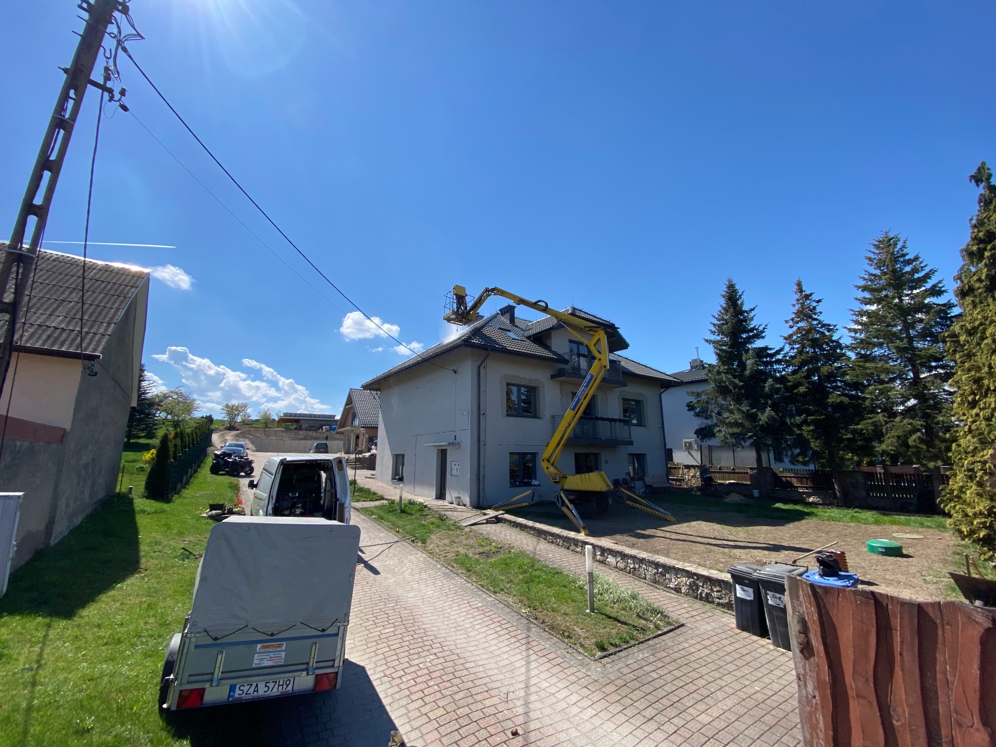 Żółty podnośnik koszowy przy szarym domu podczas mycia dachu. Widoczny fragment kostki brukowej, trawnik, drzewa iglaste i przyczepa. Słoneczny dzień.