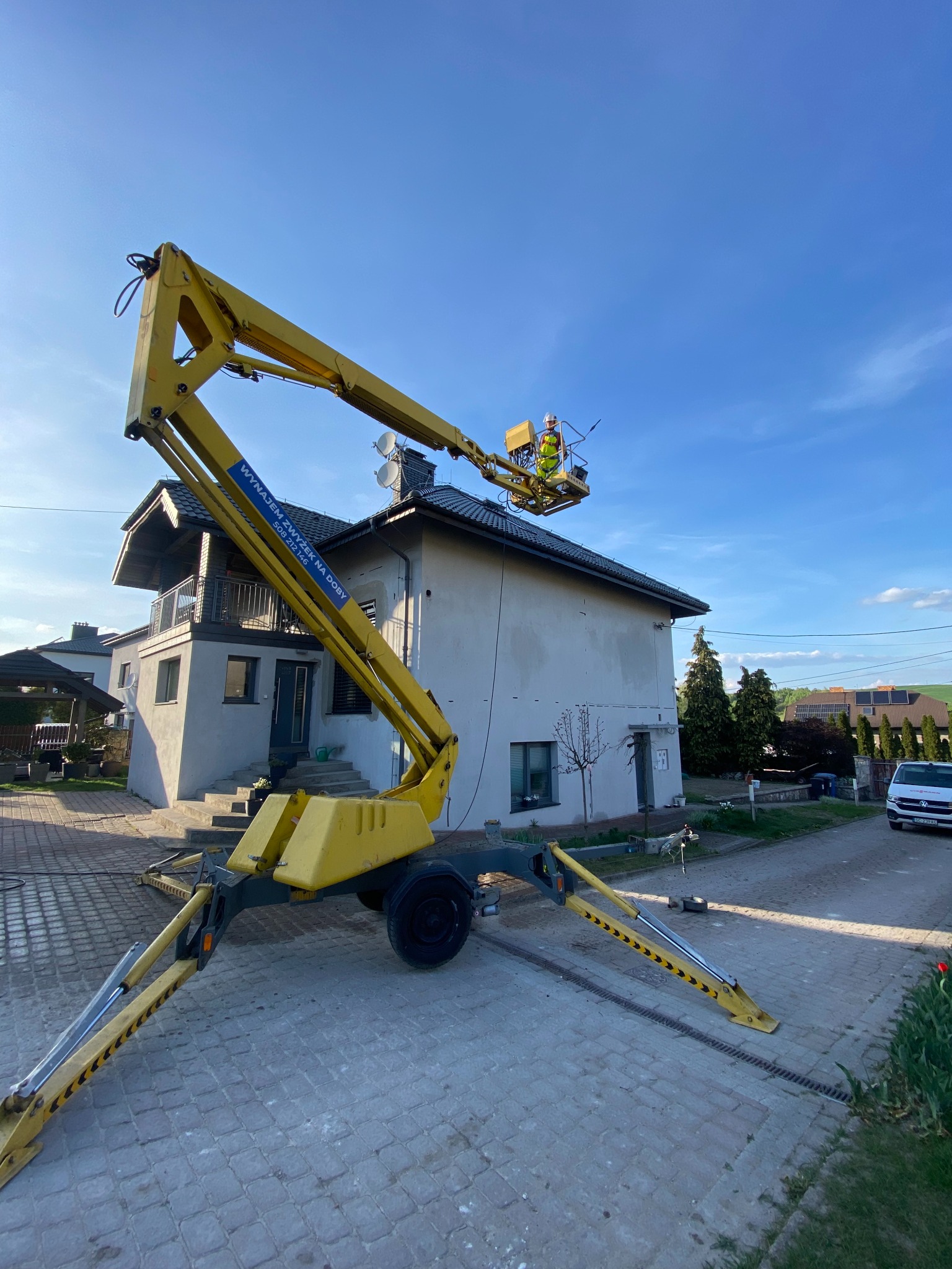 Żółty podnośnik koszowy z pracownikiem w kasku na tle jasnego domu i błękitnego nieba. Widoczne podpory podnośnika na kostce brukowej.