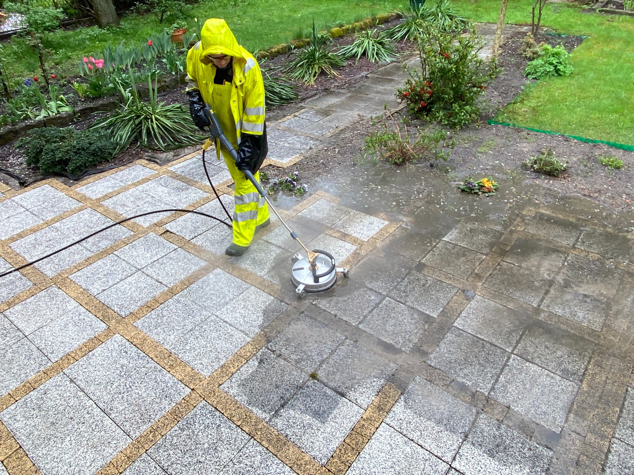 Pracownik w żółtym kombinezonie czyści kostkę brukową myjką ciśnieniową. Widoczny efekt czyszczenia na szarej kostce z beżowymi fugami, ogród w tle.