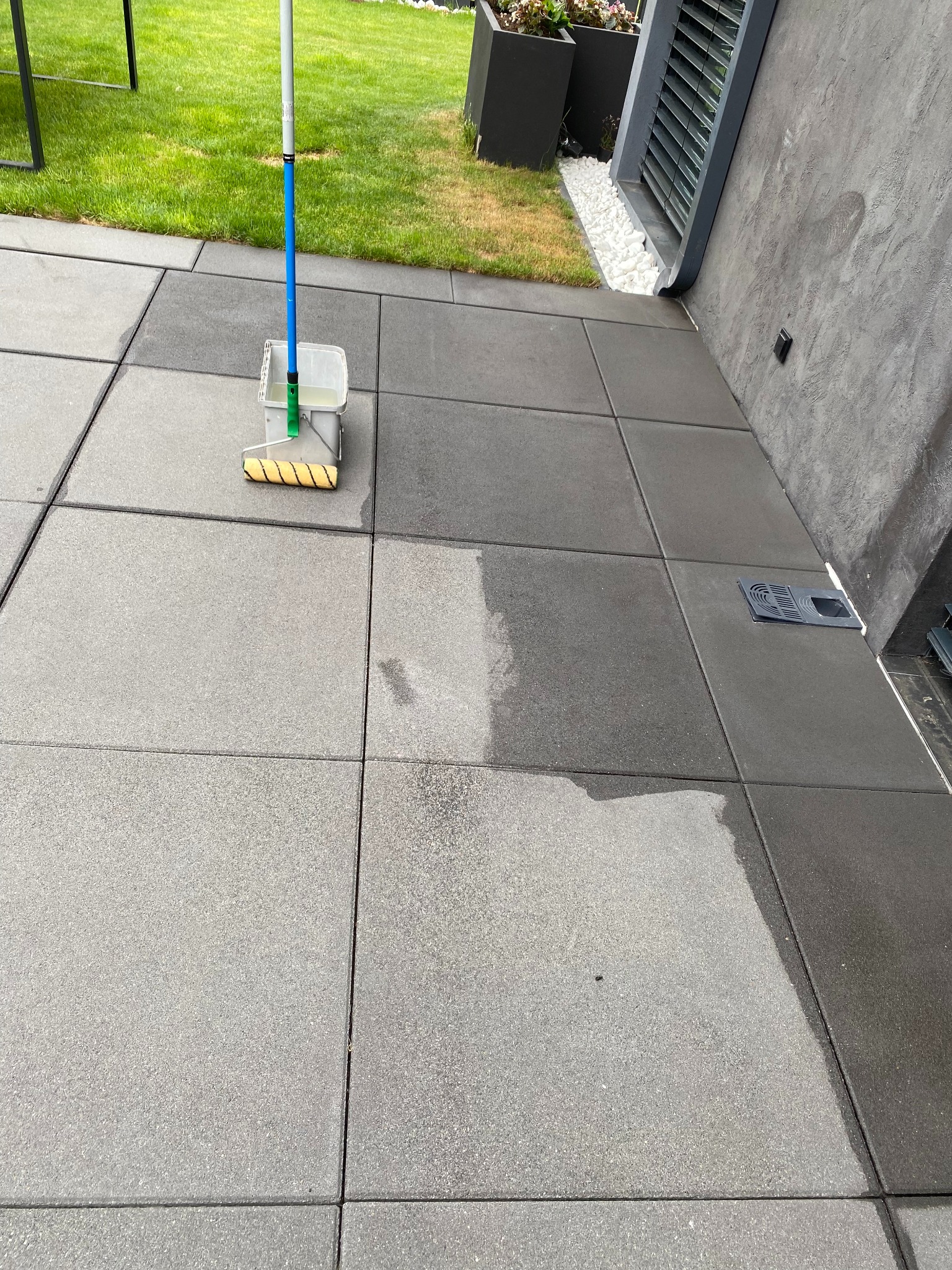 Mycie kostki brukowej: widoczna różnica między czystą a brudną powierzchnią. Wiadro z wodą i wałek do czyszczenia kostki brukowej na szarym tle.