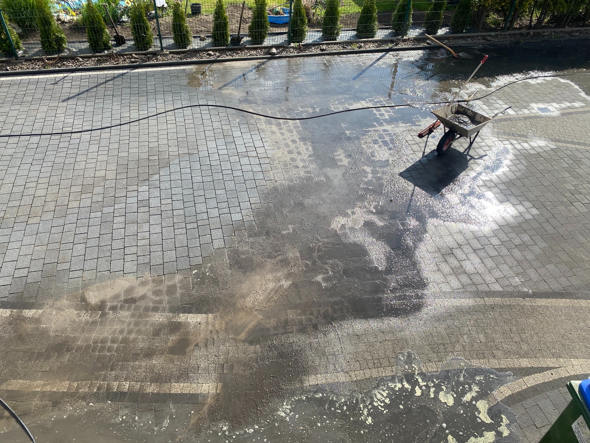 Częściowo umyta kostka brukowa z wózkiem i narzędziami w słoneczny dzień. Widoczne różnice w czystości kostki, od jasnej do ciemnej. W tle rząd tuj.