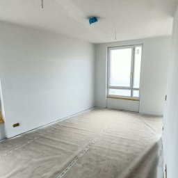 Serwis Hause - Pokój w trakcie prac wykończeniowych; ściany świeżo pomalowane na biało, podłoga zabezpieczona folią ochronną z napisem JOKA, okno zaklejone folią malarską, widoczne kable wystające z sufitu.
