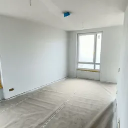 Pokój w trakcie prac wykończeniowych; ściany świeżo pomalowane na biało, podłoga zabezpieczona folią ochronną z napisem JOKA, okno zaklejone folią malarską, widoczne kable wystające z sufitu.