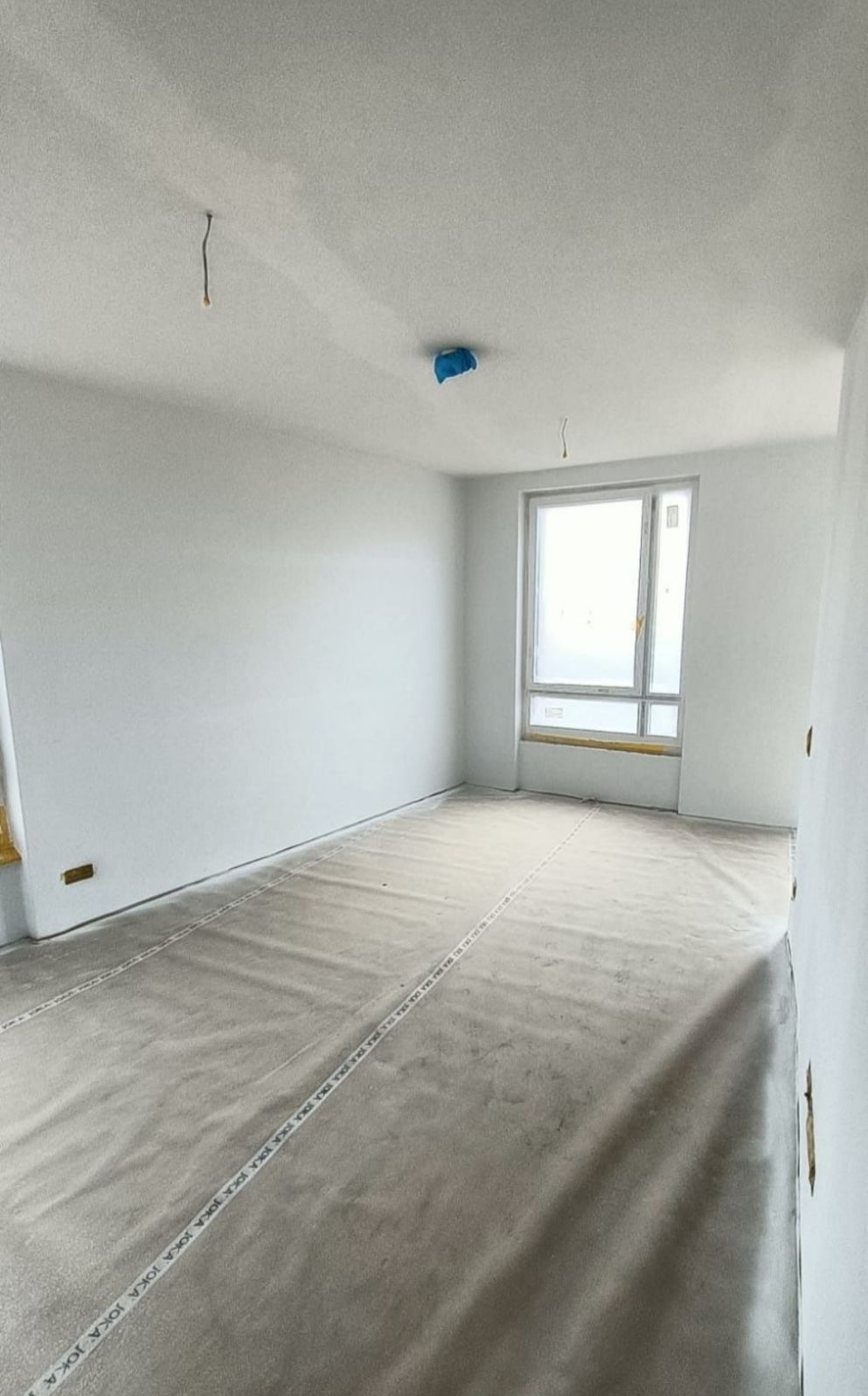 Pokój w trakcie prac wykończeniowych; ściany świeżo pomalowane na biało, podłoga zabezpieczona folią ochronną z napisem JOKA, okno zaklejone folią malarską, widoczne kable wystające z sufitu.