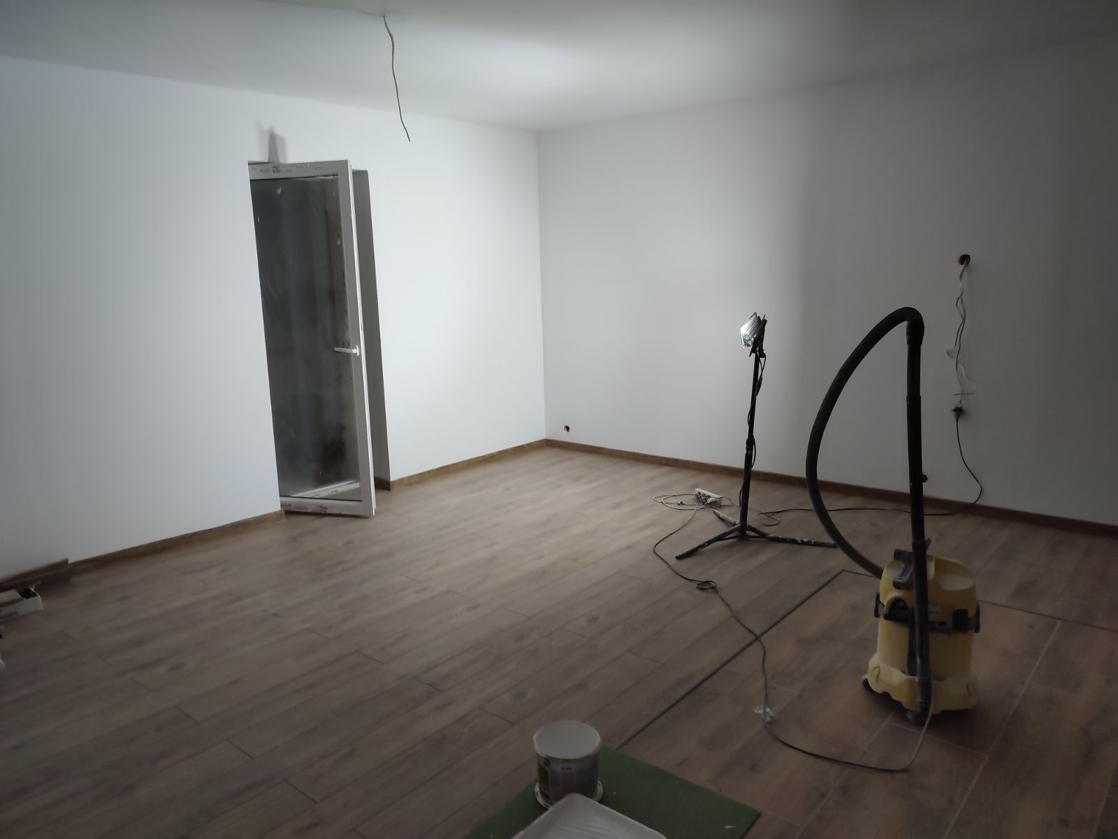 Pomalowane na biało wnętrze pokoju z drewnianą podłogą, widoczny otwarty futryna drzwi, lampa halogenowa na statywie, odkurzacz i akcesoria malarskie na podłodze.