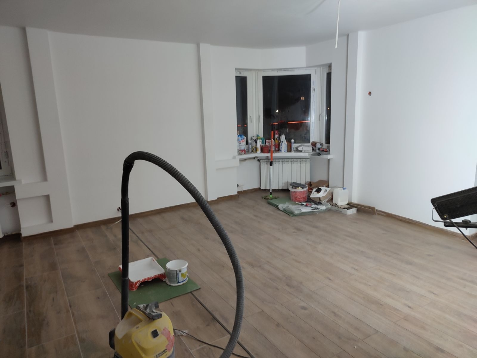 Wnętrze pokoju po wykonanych pracach przygotowawczych, ściany pomalowane na biało, widoczny odkurzacz przemysłowy, wiadro z farbą, kuweta malarska, okno z parapetem zastawionym różnymi przedmiotami.