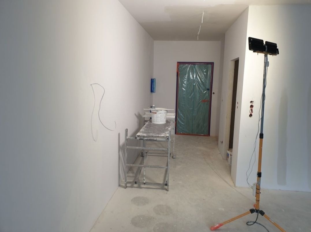Przygotowane do prac wykończeniowych wnętrze z zabezpieczonymi drzwiami folią, widoczny metalowy stół roboczy z wiadrem, szkicowany ołówek na ścianie i lampa halogenowa na statywie.