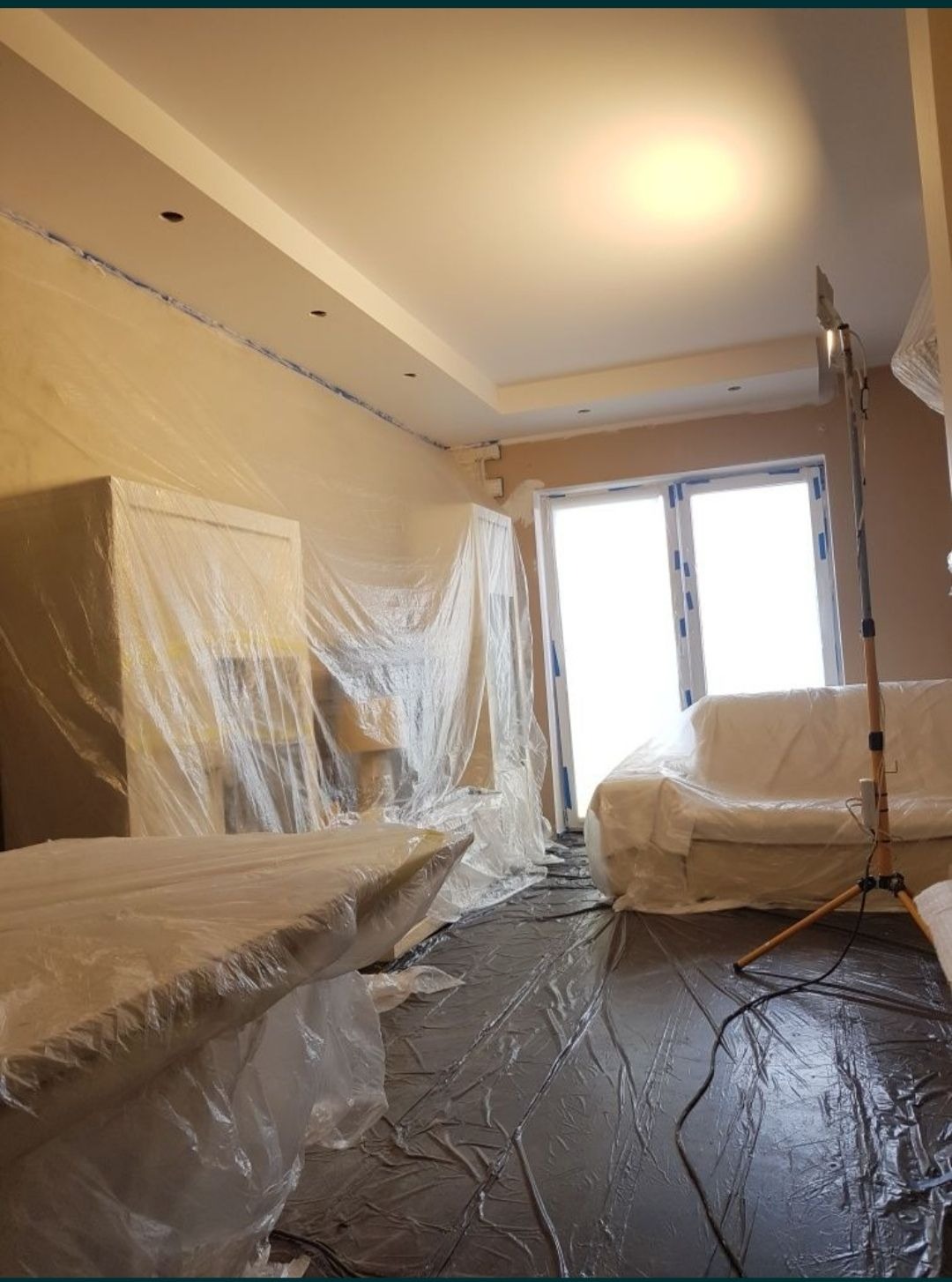 Pokój zabezpieczony folią malarską przed pracami wykończeniowymi, widoczny stół, sofa i meble okryte folią, podłoga zabezpieczona czarną folią, ściany w jasnym odcieniu beżu, sufit z oświetleniem...