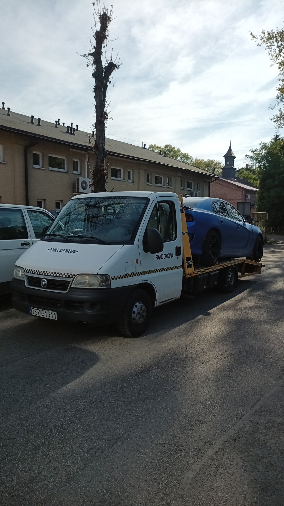 Biała laweta Fiat z napisem 'Pomoc Drogowa' przewozi niebieski samochód osobowy na tle budynku z wieżą zegarową.