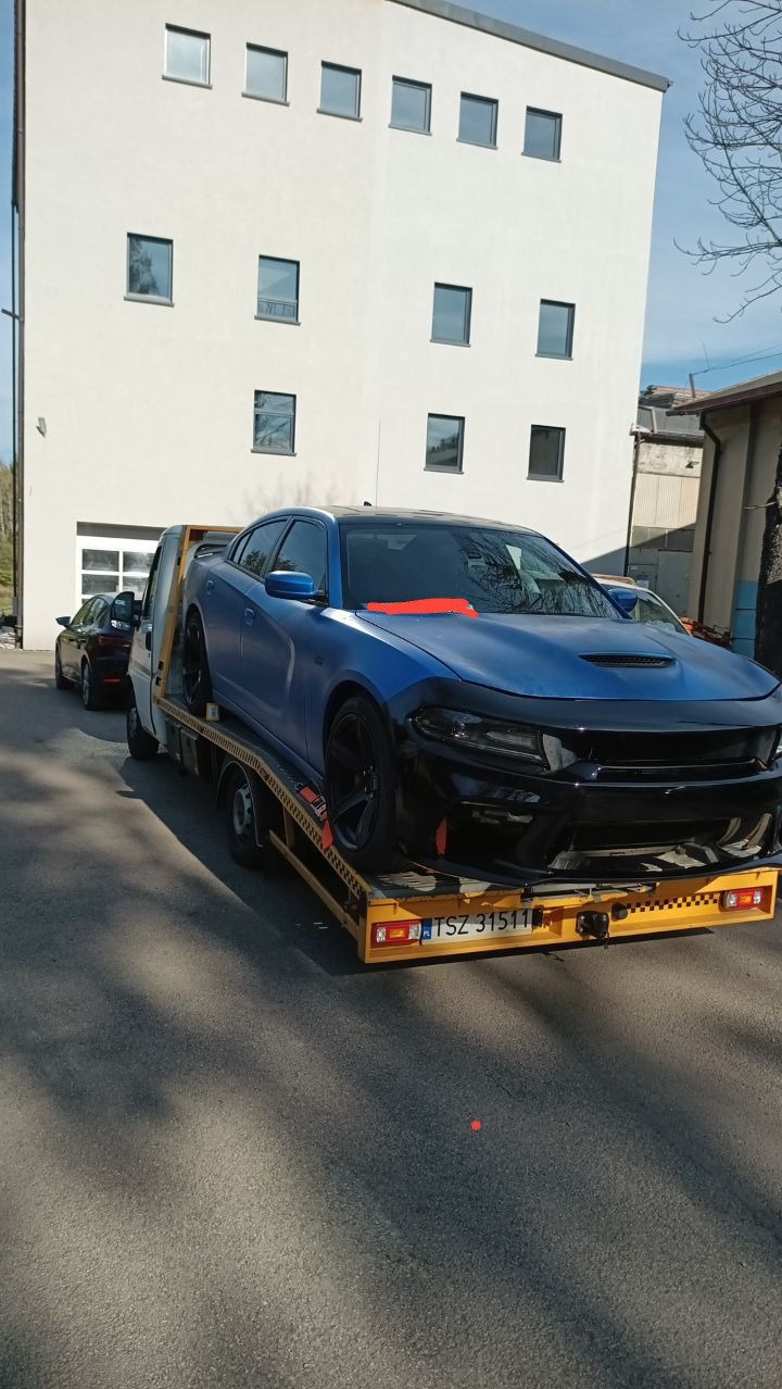 Niebieski samochód osobowy z czarnymi elementami karoserii zabezpieczony pomarańczową taśmą, transportowany na żółtej lawecie z widoczną tablicą rejestracyjną na tle jasnego budynku.