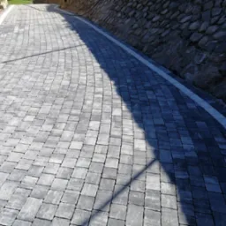 Ułożona szara kostka brukowa tworząca ścieżkę wzdłuż kamiennego muru oporowego, oddzielona białym pasem.