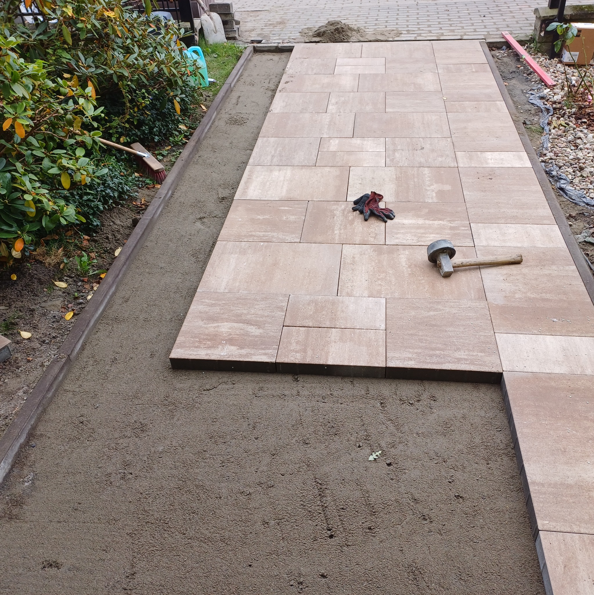 Fragment ścieżki z beżowej kostki brukowej w trakcie układania, widoczne robocze rękawice, gumowy młotek i przygotowane podłoże z piasku.