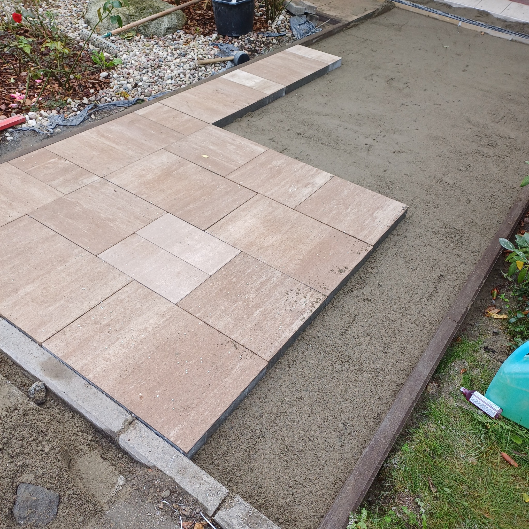 Fragment układanej powierzchni z beżowej kostki brukowej, widoczny przygotowany podkład z piasku i obrzeża, częściowo ułożone krawężniki, w tle narzędzia i roślinność.