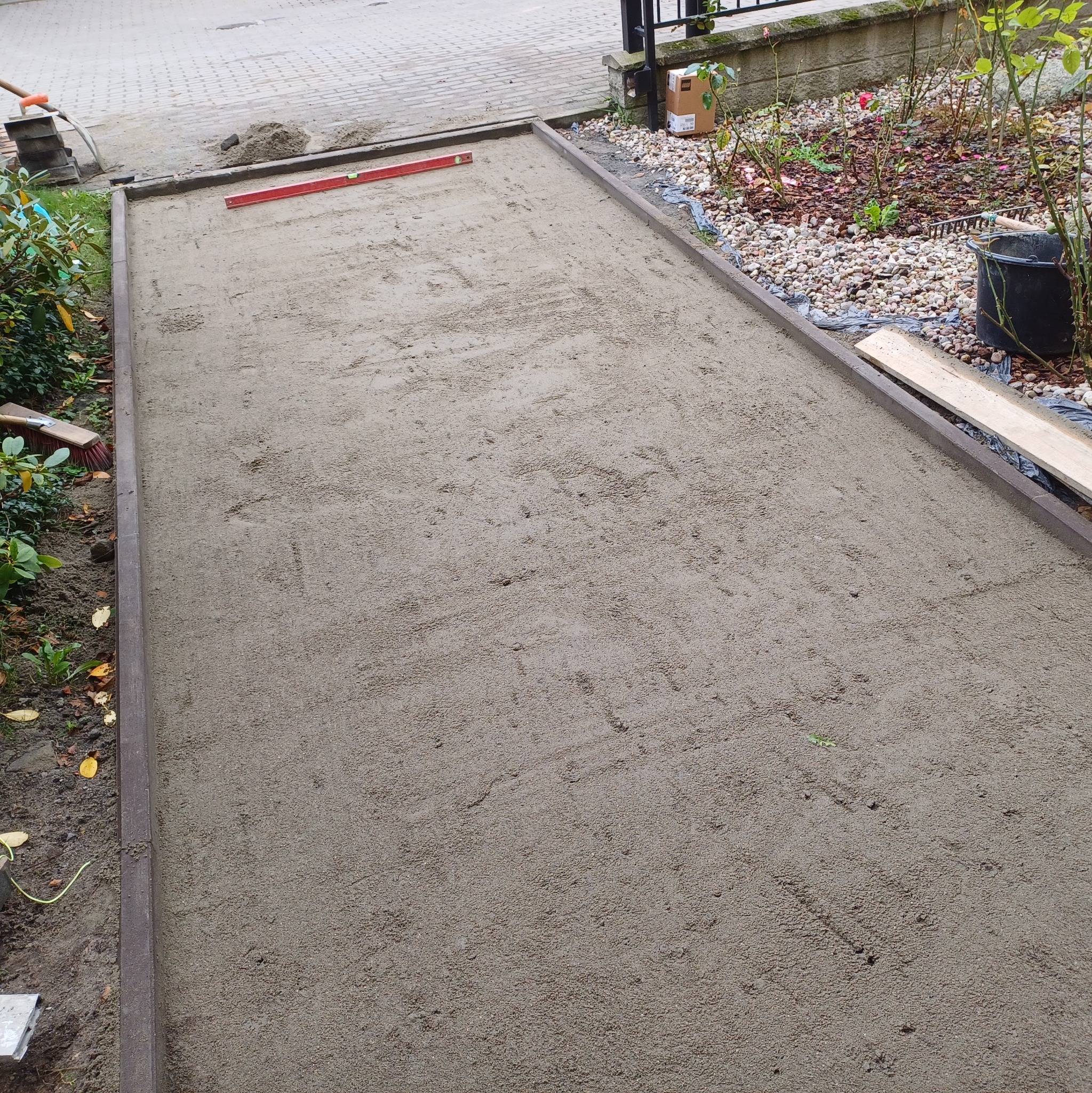 Przygotowany podkład z piasku pod przyszłą nawierzchnię, obramowany obrzeżami, z poziomicą w rogu, widoczny sprzęt budowlany i roślinność w tle.