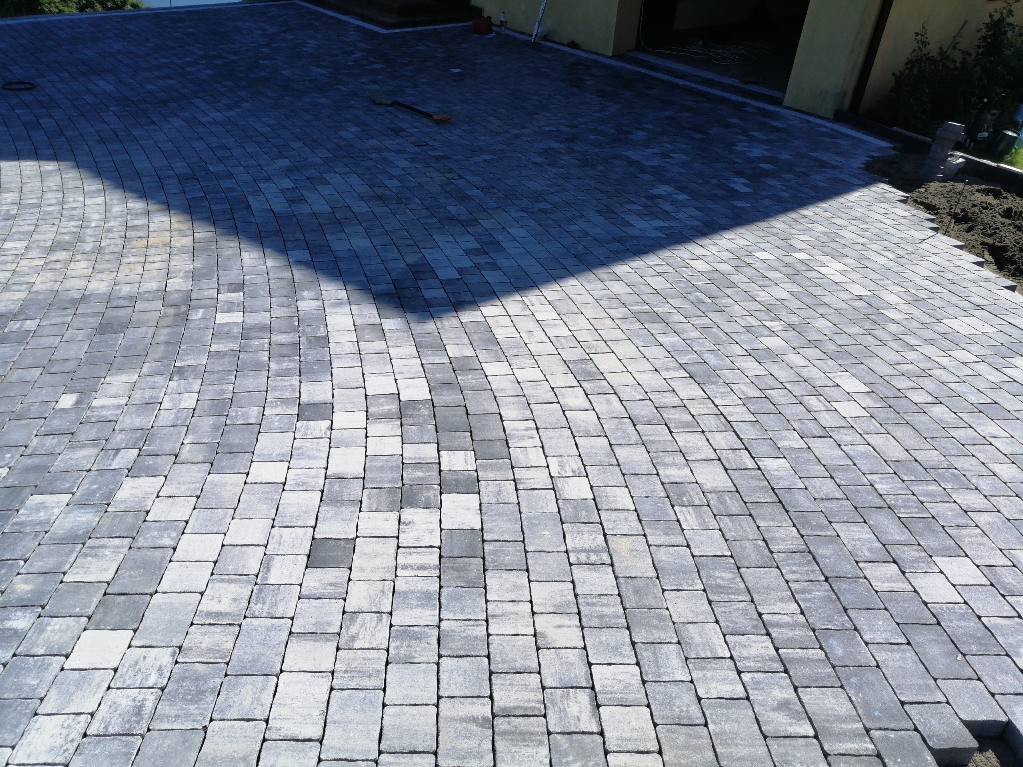 Nowa nawierzchnia z szarej kostki brukowej ułożonej w jodełkę na podjeździe, widoczne świeże obsypanie ziemią przy krawędzi, cień części budynku pada na fragment powierzchni.