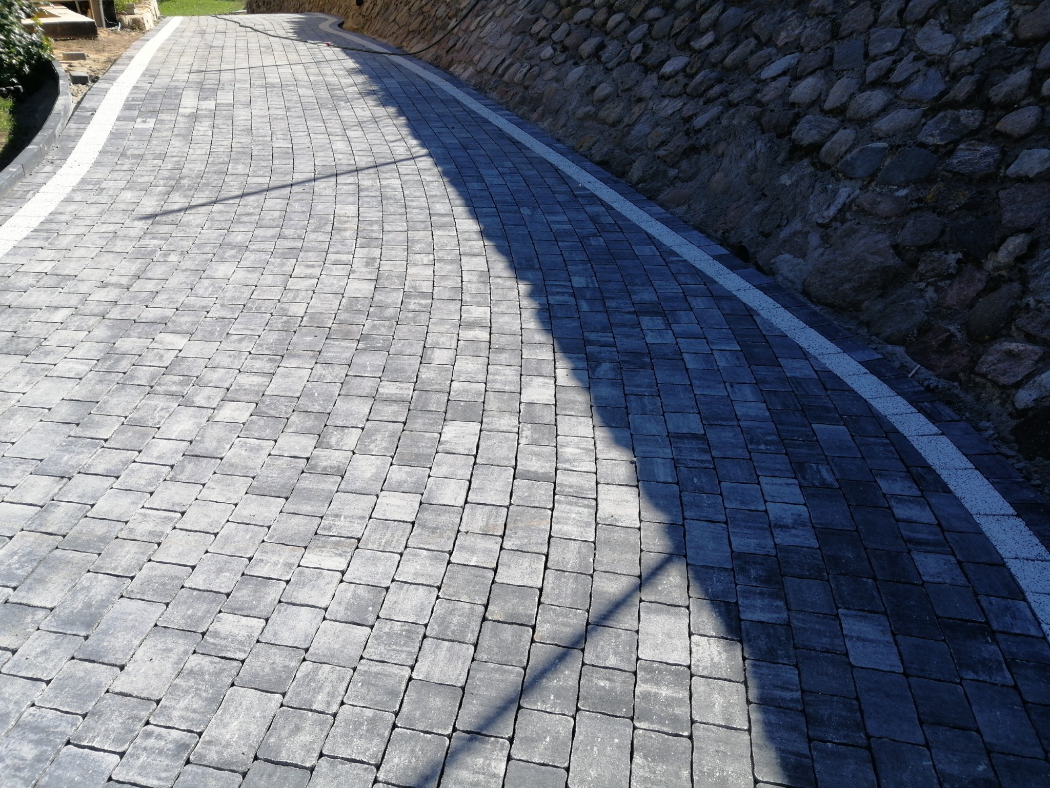 Ułożona szara kostka brukowa tworząca ścieżkę wzdłuż kamiennego muru oporowego, oddzielona białym pasem.