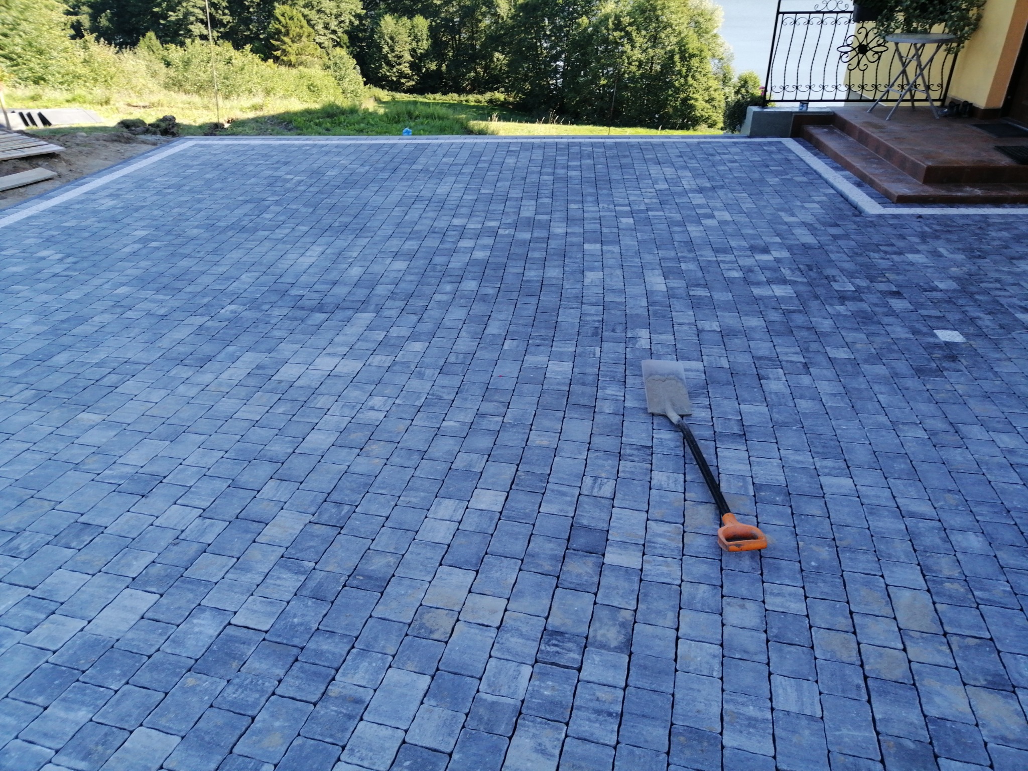 Świeżo ułożona, szara kostka brukowa na podjeździe, z widoczną łopatą z pomarańczową rączką leżącą na powierzchni, obok domu z widocznym fragmentem schodów i metalowej balustrady.