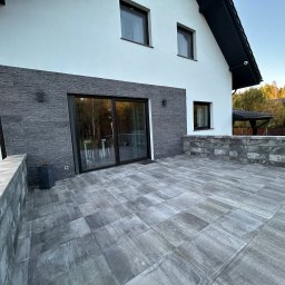 Bruinvest Tomasz Brucki - Nowoczesny taras z szarych płyt betonowych, przylegający do białego domu z elementami okładziny z szarego kamienia, widoczne przesuwne drzwi tarasowe i fragment ogrodu w tle.