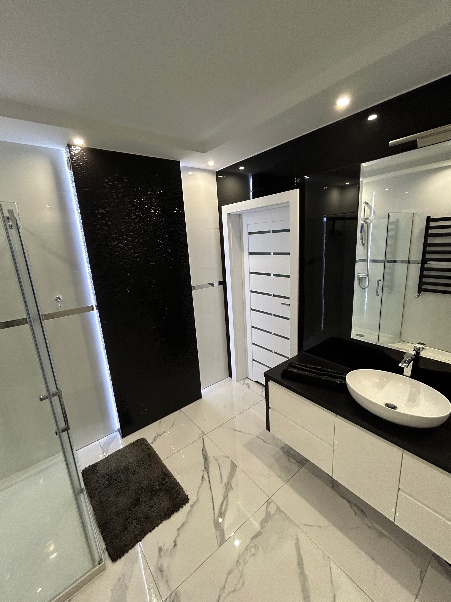 Nowoczesna łazienka z czarno-białymi płytkami marmurowymi, kabiną prysznicową ze szklanymi drzwiami, umywalką nablatową w owalnym kształcie i białymi drzwiami z poziomymi przeszkleniami.