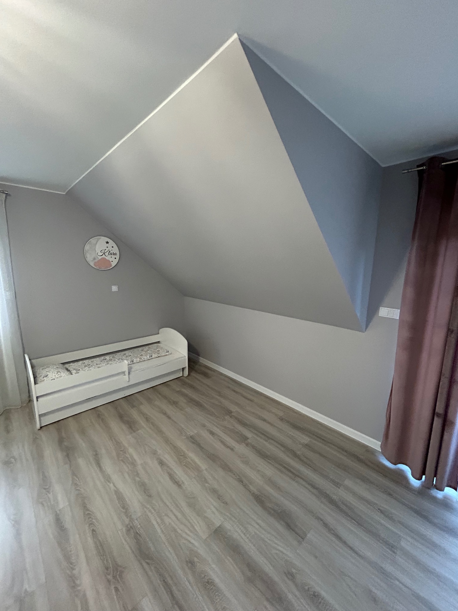 Pokój dziecięcy na poddaszu z drewnopodobną podłogą, białym łóżeczkiem i szarą ścianą z dekoracyjnym zegarem z imieniem Klara.