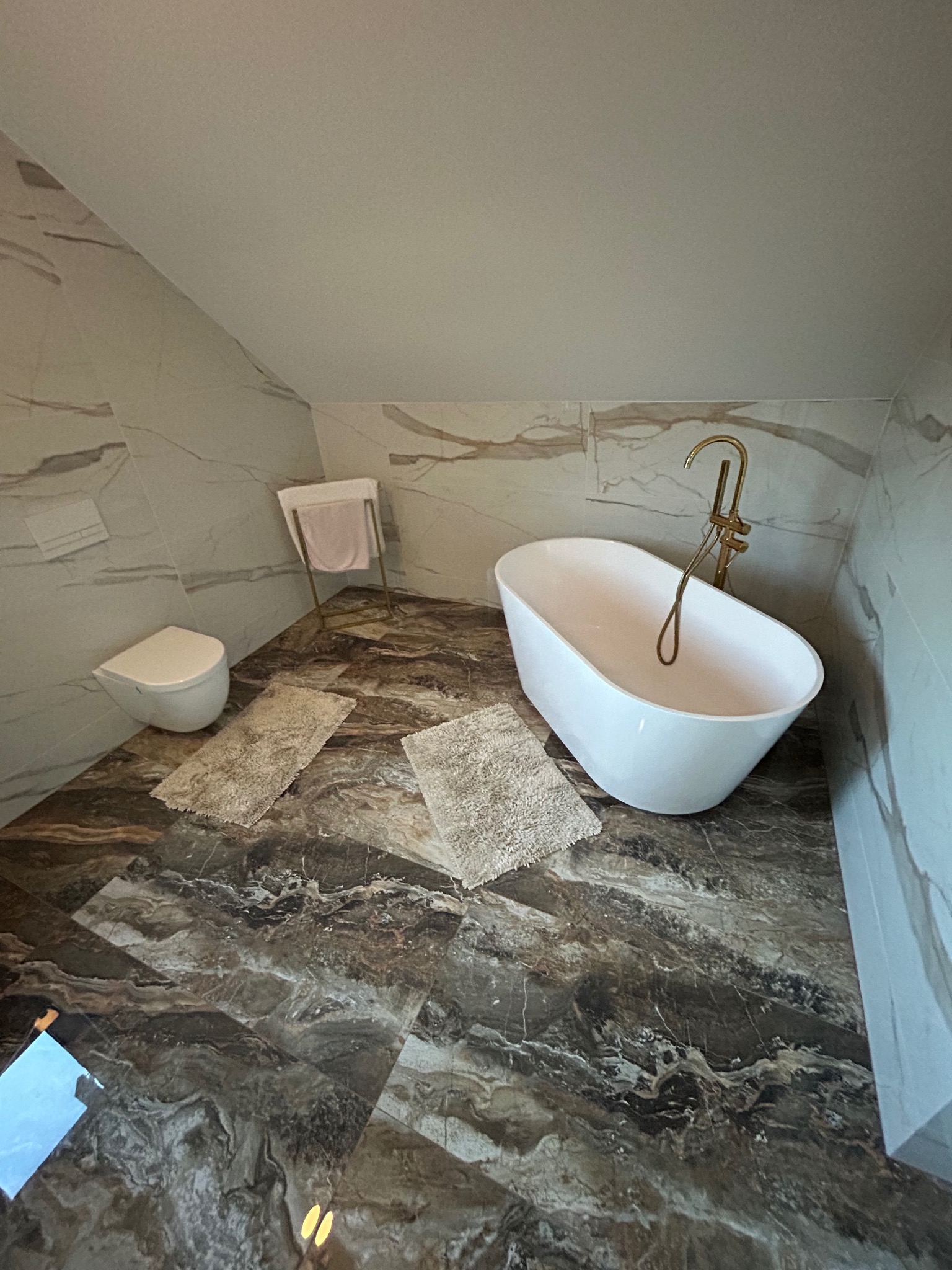 Nowoczesna łazienka na poddaszu z wolnostojącą wanną, marmurowymi płytkami na ścianach i podłodze, oraz złotą armaturą, oświetlona naturalnym światłem wpadającym z okna odbijającym się w podłodze.