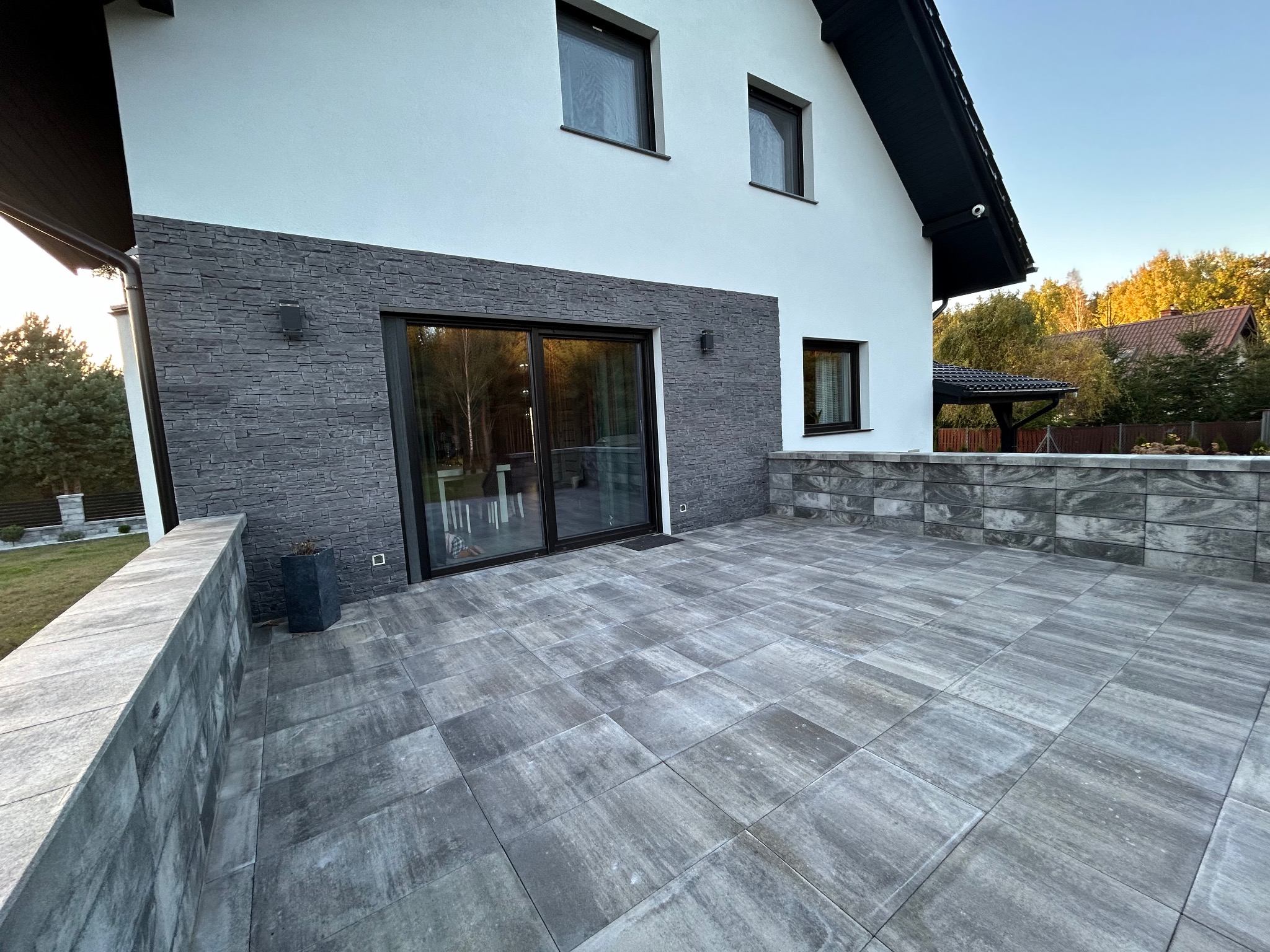 Nowoczesny taras z szarych płyt betonowych, przylegający do białego domu z elementami okładziny z szarego kamienia, widoczne przesuwne drzwi tarasowe i fragment ogrodu w tle.