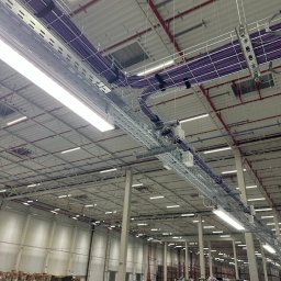 KBCS ELEKTRO-TECHNIKA - Instalacja elektryczna w hali magazynowej: widok na koryta kablowe z fioletowymi kablami pod sufitem, oświetlenie LED, stalowe słupy konstrukcyjne i regały magazynowe w tle.
