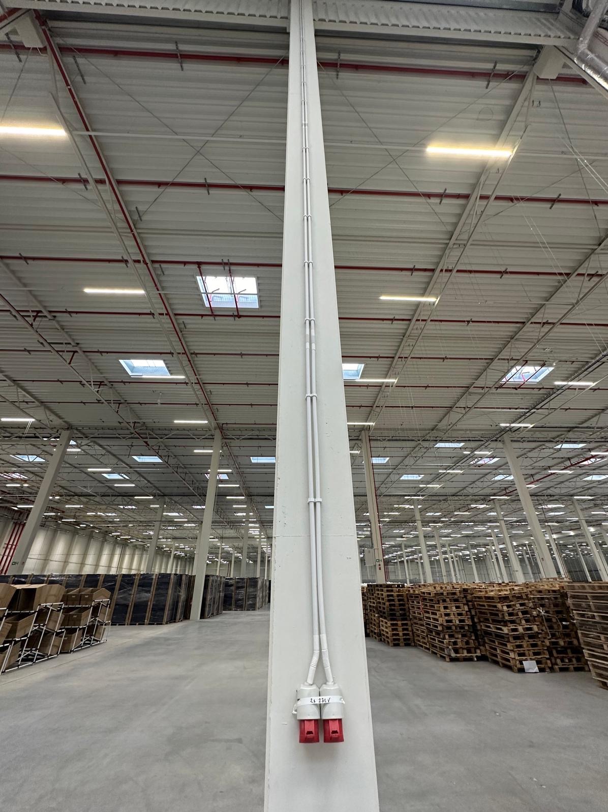 Dwa czerwone gniazda siłowe 230V na betonowym słupie w hali magazynowej z widocznymi rurami instalacyjnymi, oświetleniem i paletami w tle.